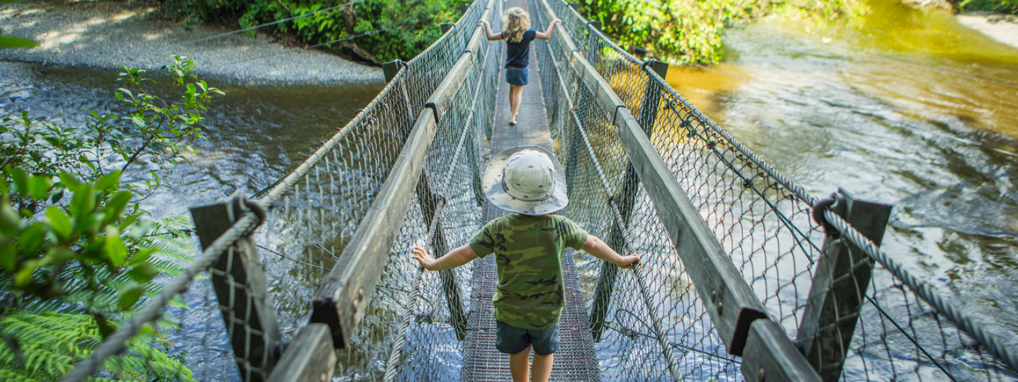 Two kids running along a suspension bridge over a river and surrounded by plants and trees.